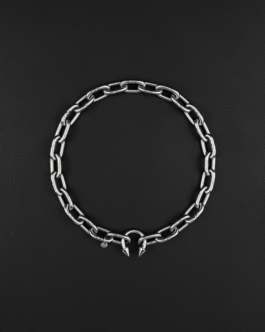 stainless steel Silver chunky heavy thick chain necklace. The necklace is composed of linked segments and a piercing barbel clasp with two spiked ends. The jewellery is set against a dark backdrop.