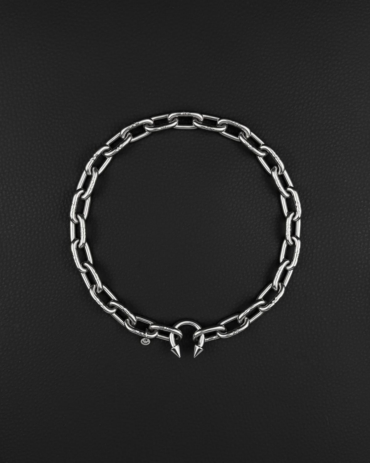 stainless steel Silver chunky heavy thick chain necklace. The necklace is composed of linked segments and a piercing barbel clasp with two spiked ends. The jewellery is set against a dark backdrop.