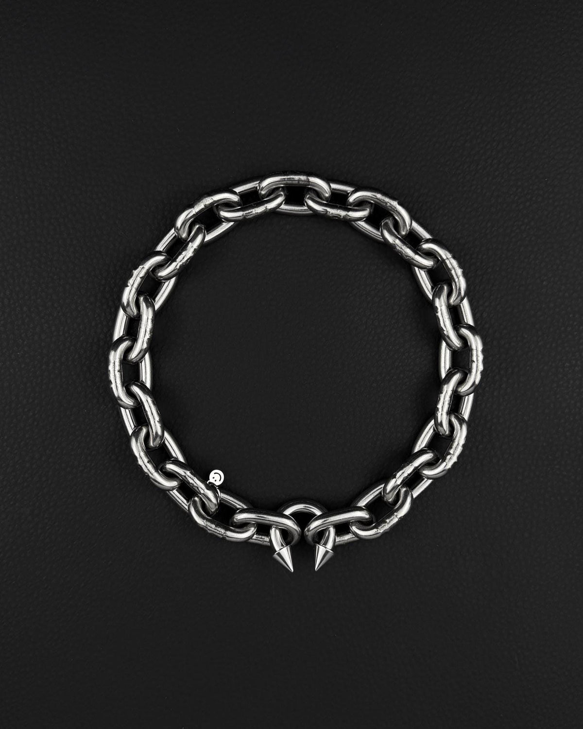 A stainless steel silver chunky heavy thick industrial chain necklace. The necklace has two pointed spikes at the clasp, the clasp is a piercing barbel. The jewellery is displayed on a black surface.