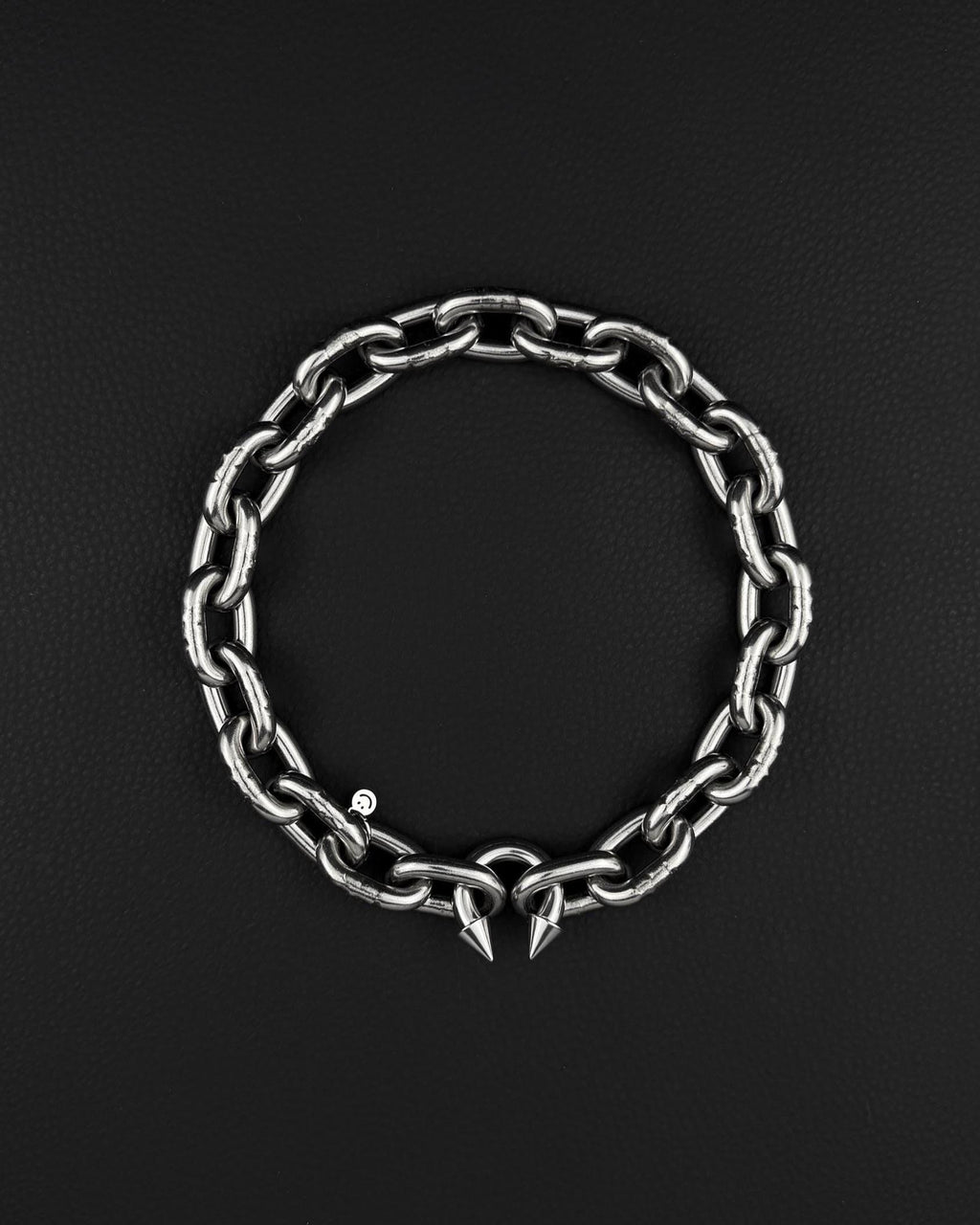 A stainless steel silver chunky heavy thick industrial chain necklace. The necklace has two pointed spikes at the clasp, the clasp is a piercing barbel. The jewellery is displayed on a black surface.