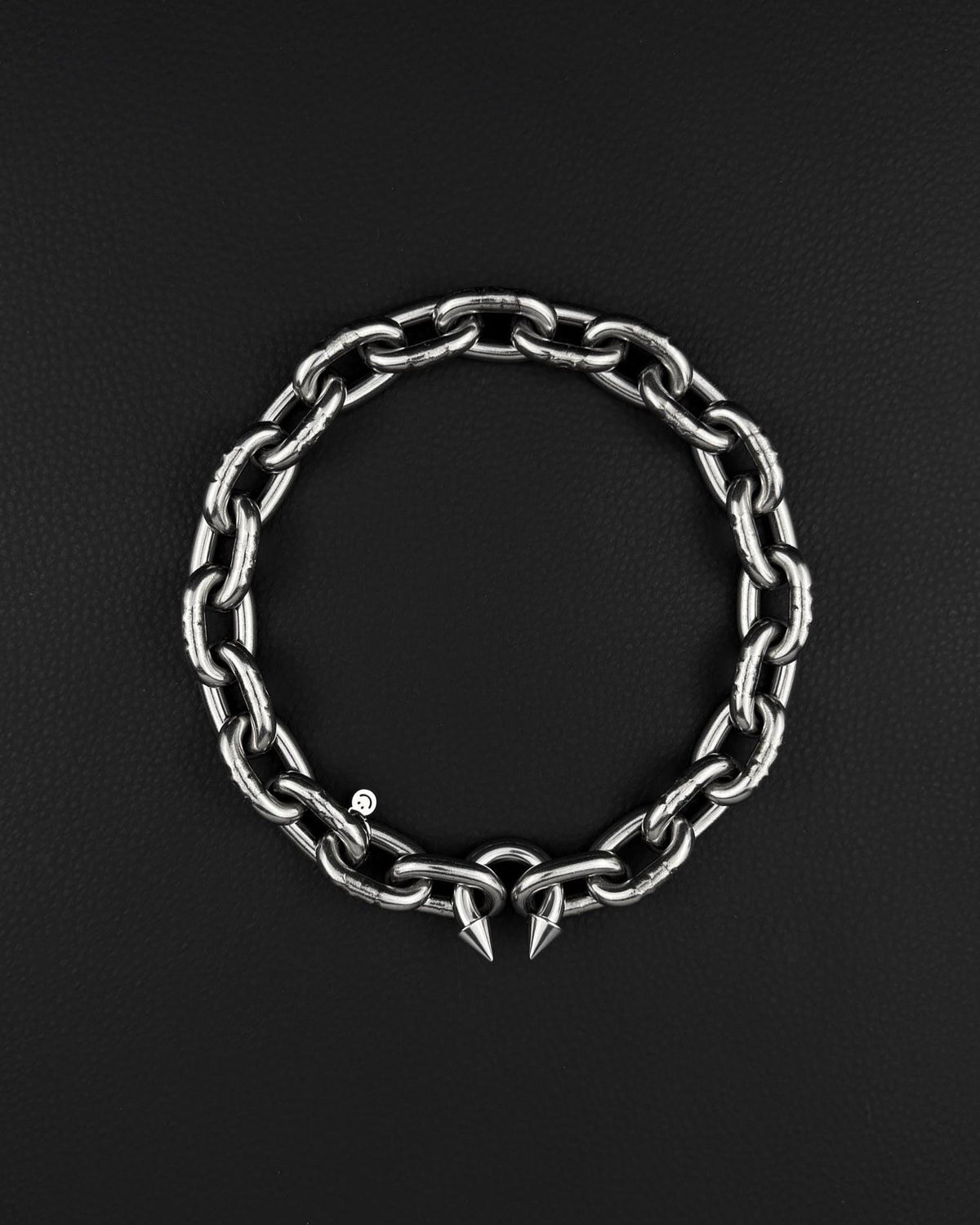 A stainless steel silver chunky heavy thick industrial chain necklace. The necklace has two pointed spikes at the clasp, the clasp is a piercing barbel. The jewellery is displayed on a black surface.