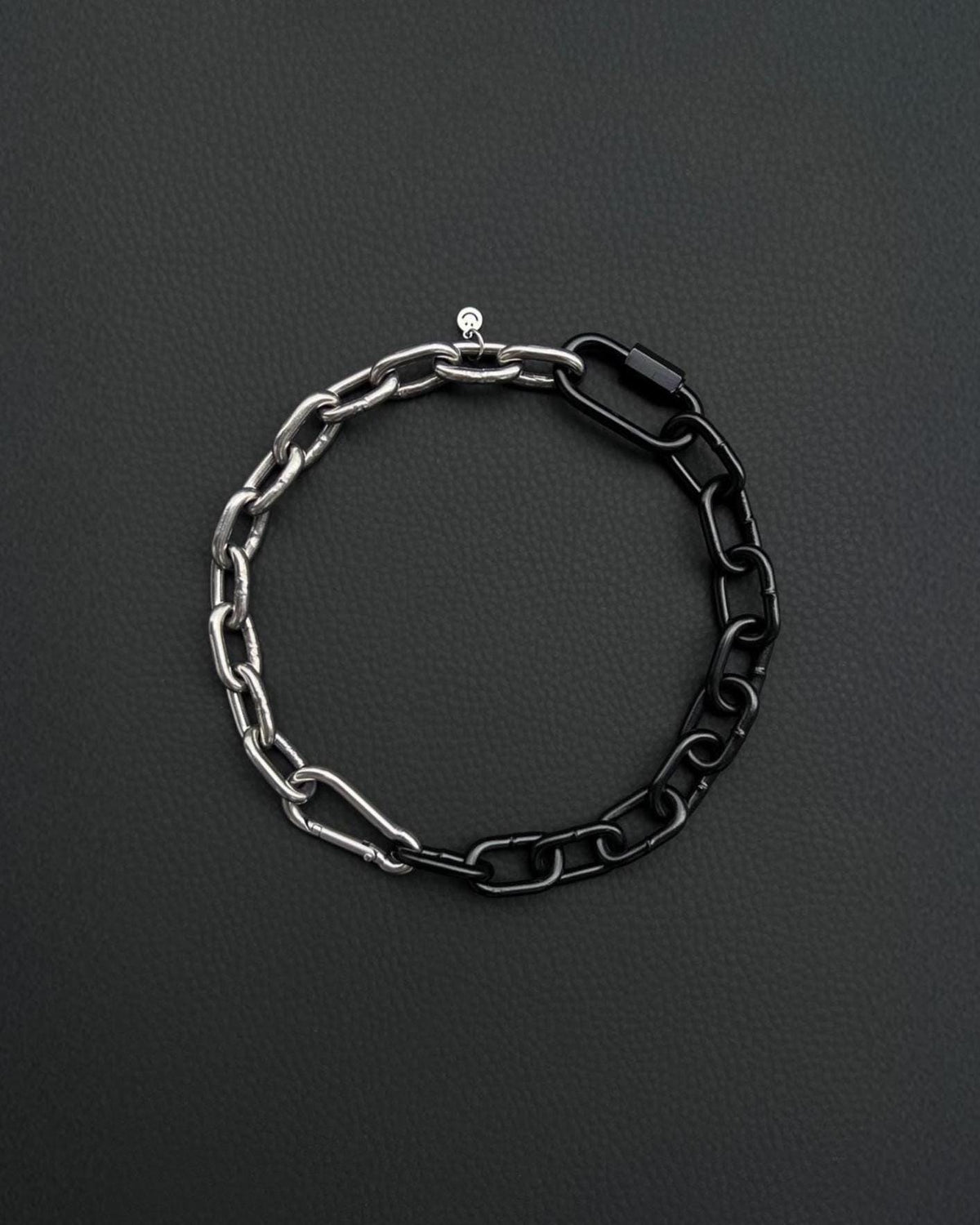 A two-tone stainless steel metal chunky heavy chain necklace. One half is silver, the other black. The necklace is made of interlocking chain links and has a small clasp. The background is a dark, textured surface.
