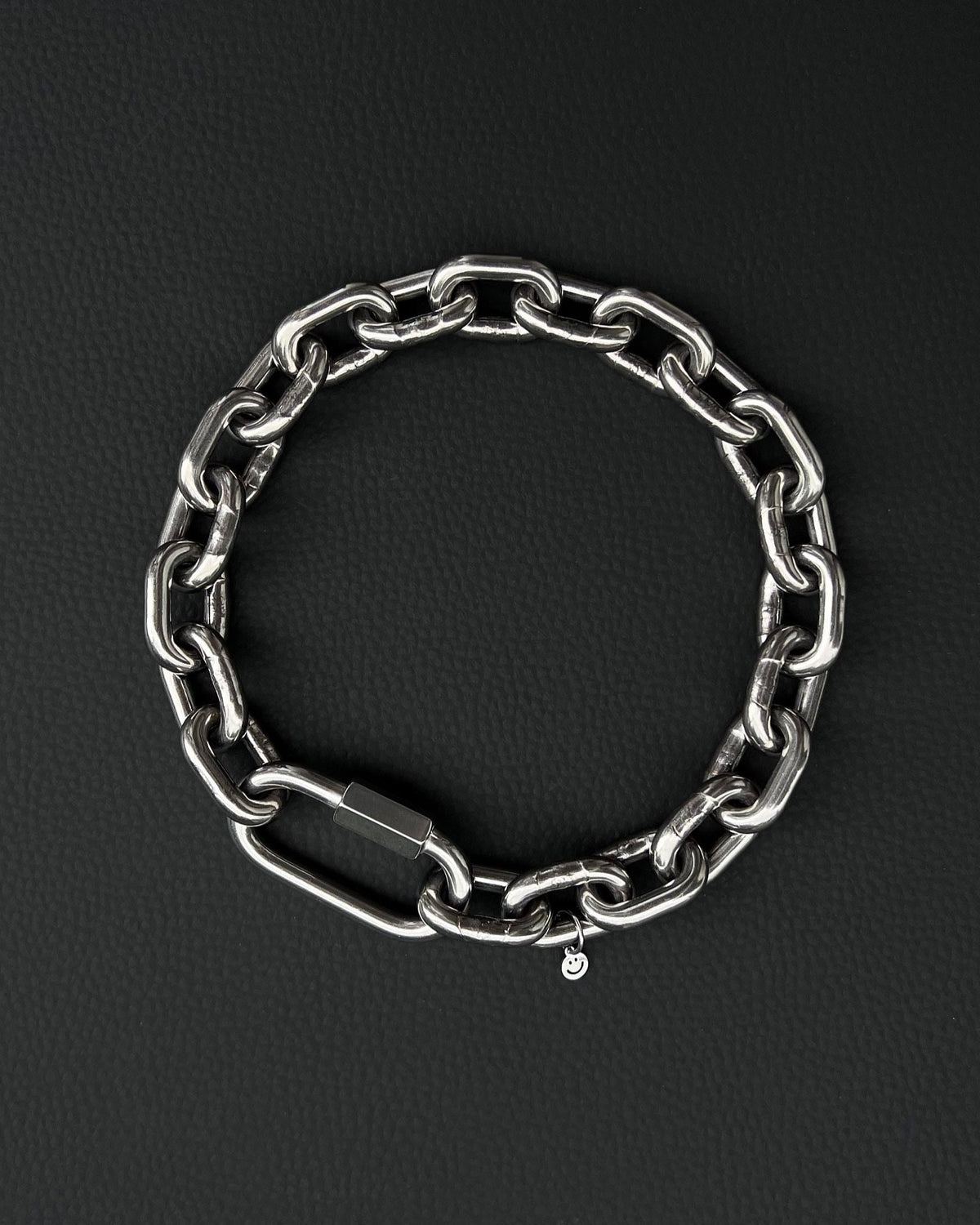 A thick silver stainless steel chain necklace with a large carabiner clasp. The choker is made of thick, oversized, interlocking links and has a small charm with a smiley face. The necklace is displayed on a black surface. the chain is made of 6mm thick links. queer aesthetics