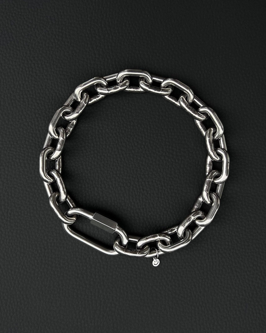 A thick silver stainless steel chain necklace with a large carabiner clasp. The choker is made of thick, oversized, interlocking links and has a small charm with a smiley face. The necklace is displayed on a black surface.