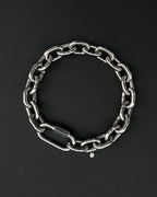 A thick silver stainless steel chain necklace with a large carabiner clasp. The choker is made of thick, oversized, interlocking links and has a small charm with a smiley face. The necklace is displayed on a black surface.