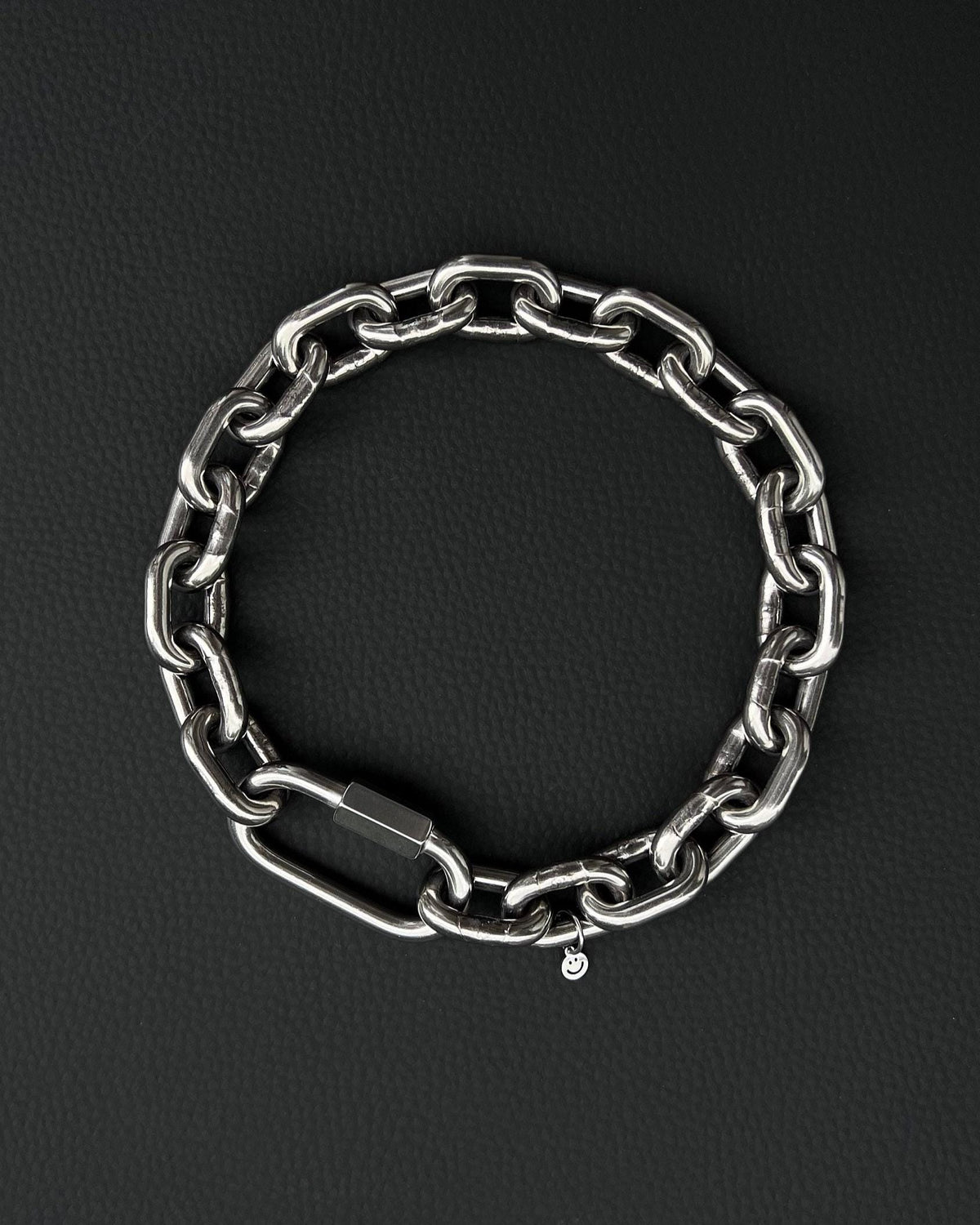 A thick silver stainless steel chain necklace with a large carabiner clasp. The choker is made of thick, oversized, heavy interlocking links and has a small charm with a smiley face. The necklace is displayed on a black surface. the chain is made of 6mm thick links. queer aesthetics