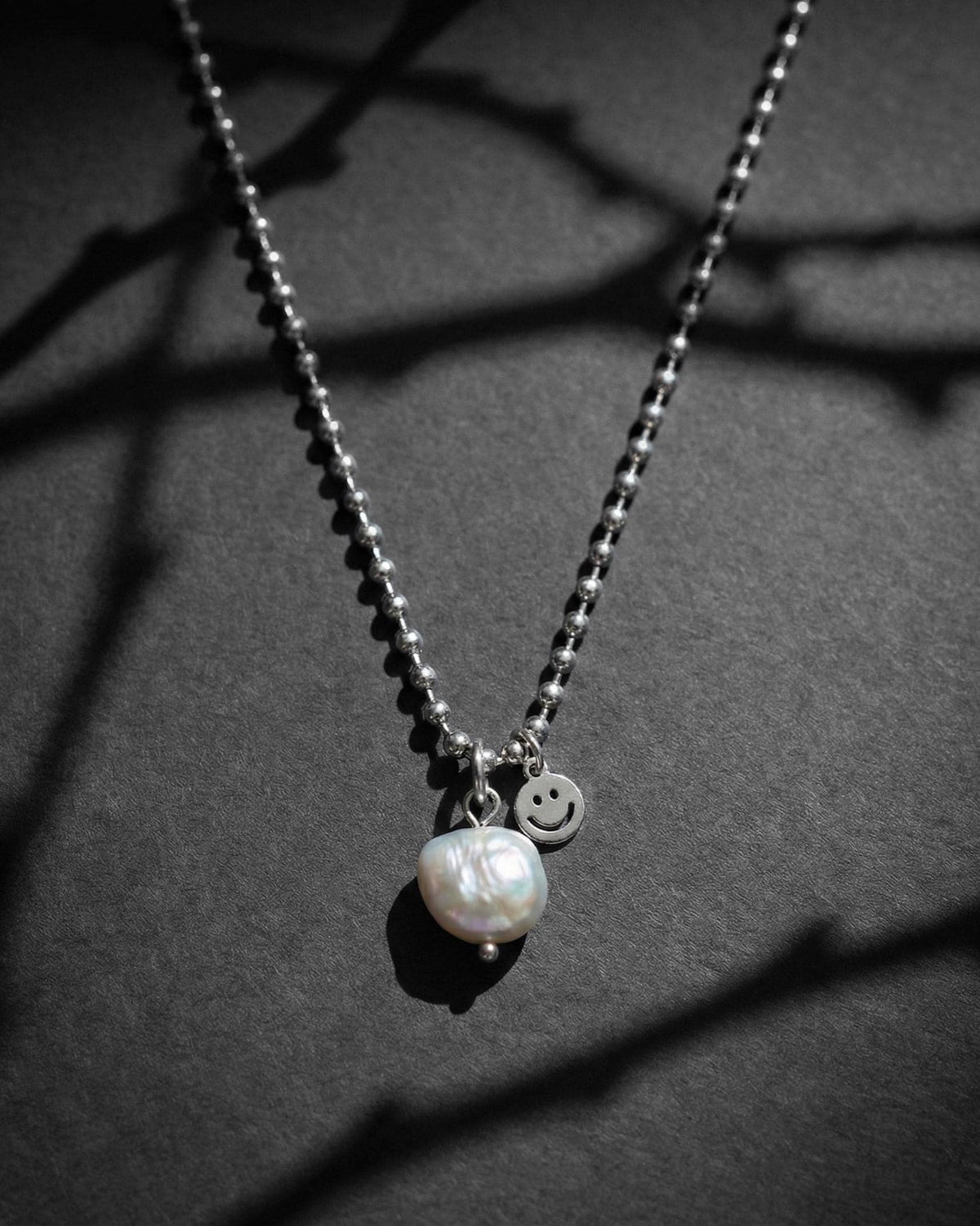 A silver-toned stainless steel chain necklace featuring a real baroque white iridescent grey pearl pendant and a small circular charm with an engraved smiley face. The necklace has a ball chain design, and the pendant is attached with a simple silver jump ring.