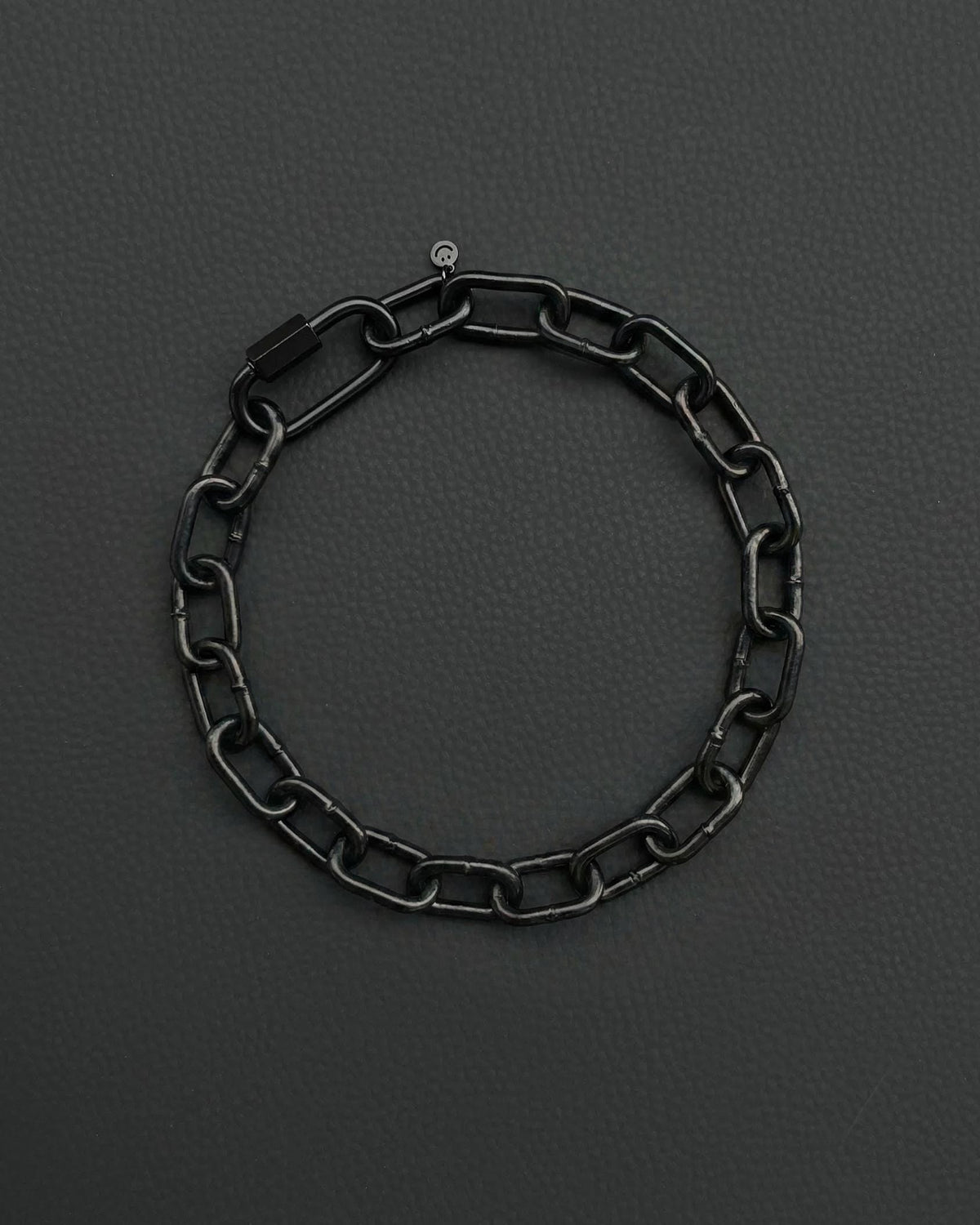 A black chunky heavy chain necklace with a rectangular clasp and a small circular charm. The bracelet is made of interlocking oval links and is set against a dark grey background.