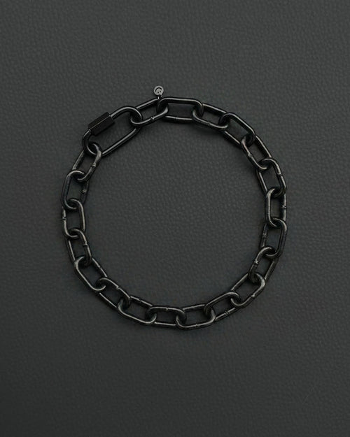 A black chunky heavy chain necklace with a rectangular clasp and a small circular charm. The bracelet is made of interlocking oval links and is set against a dark grey background.
