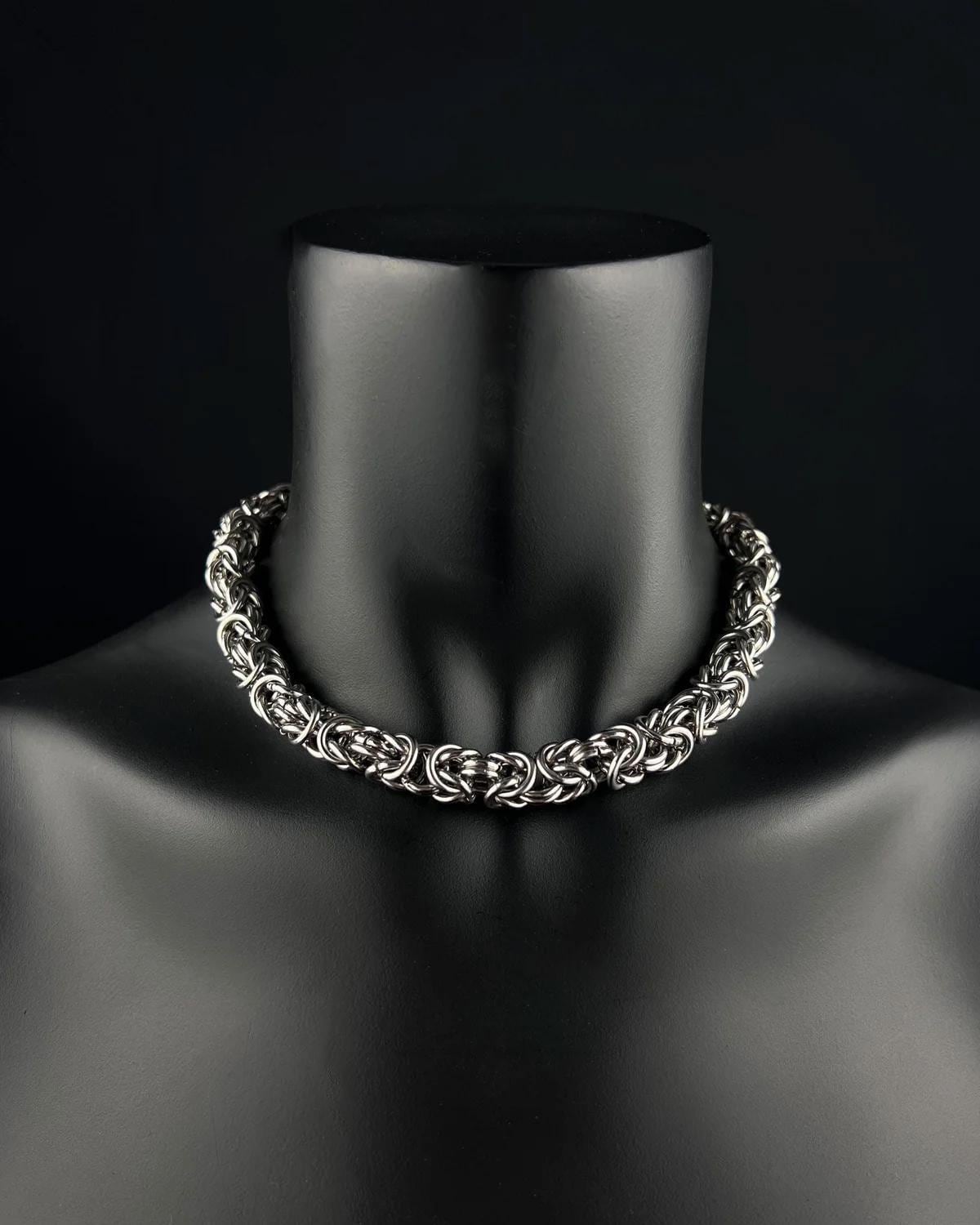 A stainless steel silver Byzantine chainmaille chunky chain necklace on a black mannequin. The necklace is made of interwoven metal rings, creating a thick, textured appearance. The background is a solid black.