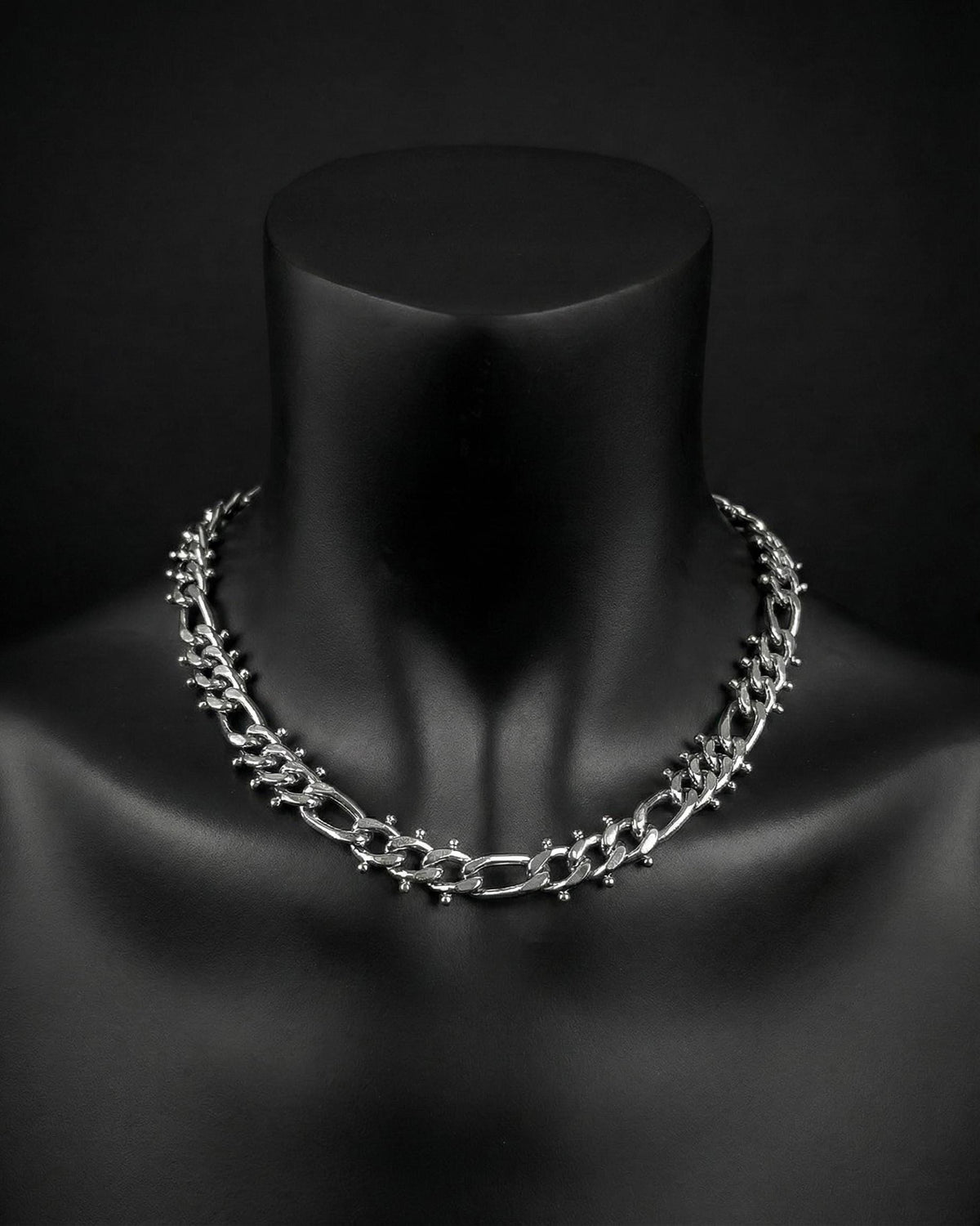 A stainless steel silver chain necklace with a chunky, textured design. The necklace is displayed on a black mannequin. The chain links are thick and feature small, decorative studs. The overall aesthetic is bold and modern.