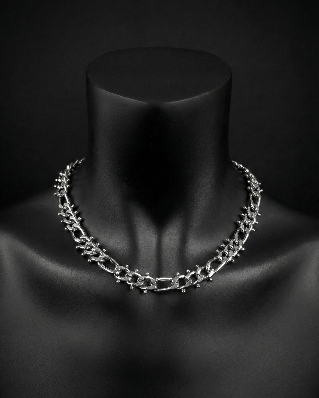 A stainless steel silver chain necklace with a chunky, textured design. The necklace is displayed on a black mannequin. The chain links are thick and feature small, decorative studs. The overall aesthetic is bold and modern.