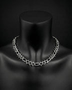 A stainless steel silver chain necklace with a chunky, textured design. The necklace is displayed on a black mannequin. The chain links are thick and feature small, decorative studs. The overall aesthetic is bold and modern.