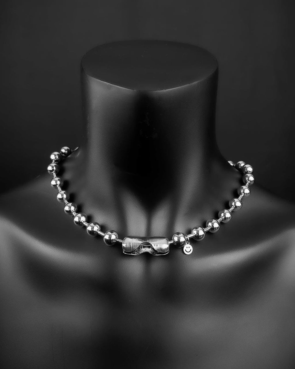 A stainless steel silver necklace with a ball chain design. The necklace has a small charm with a smiley face. The jewellery is displayed on a black mannequin.