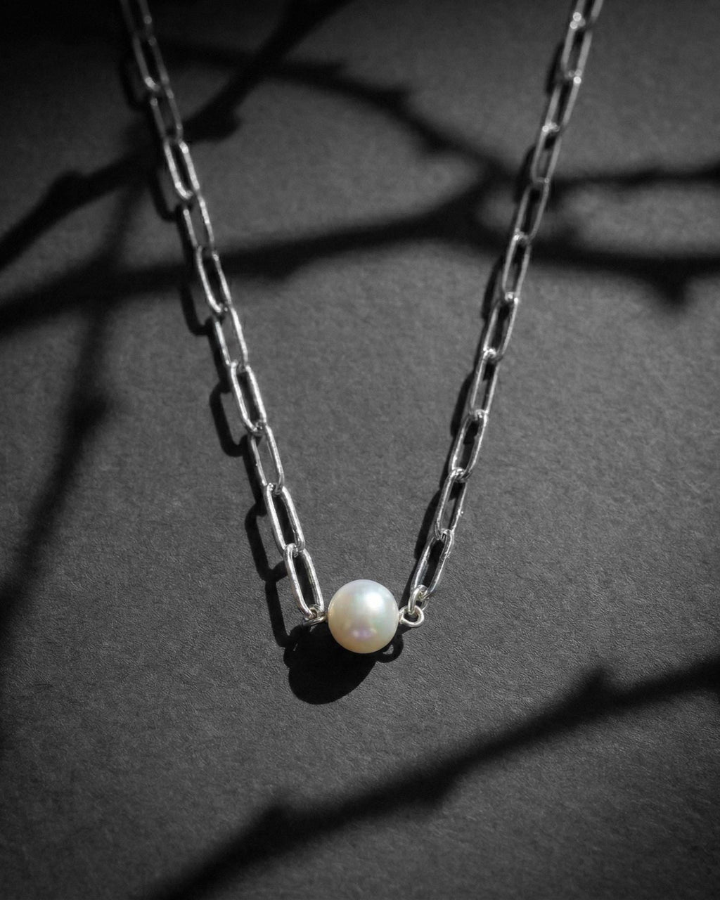 A silver-toned stainless steel chain necklace featuring a real small white pearl pendant and a small circular charm with an engraved smiley face. The necklace has a paper clip -style link chain design, and the pendant is attached with a simple silver jump ring.