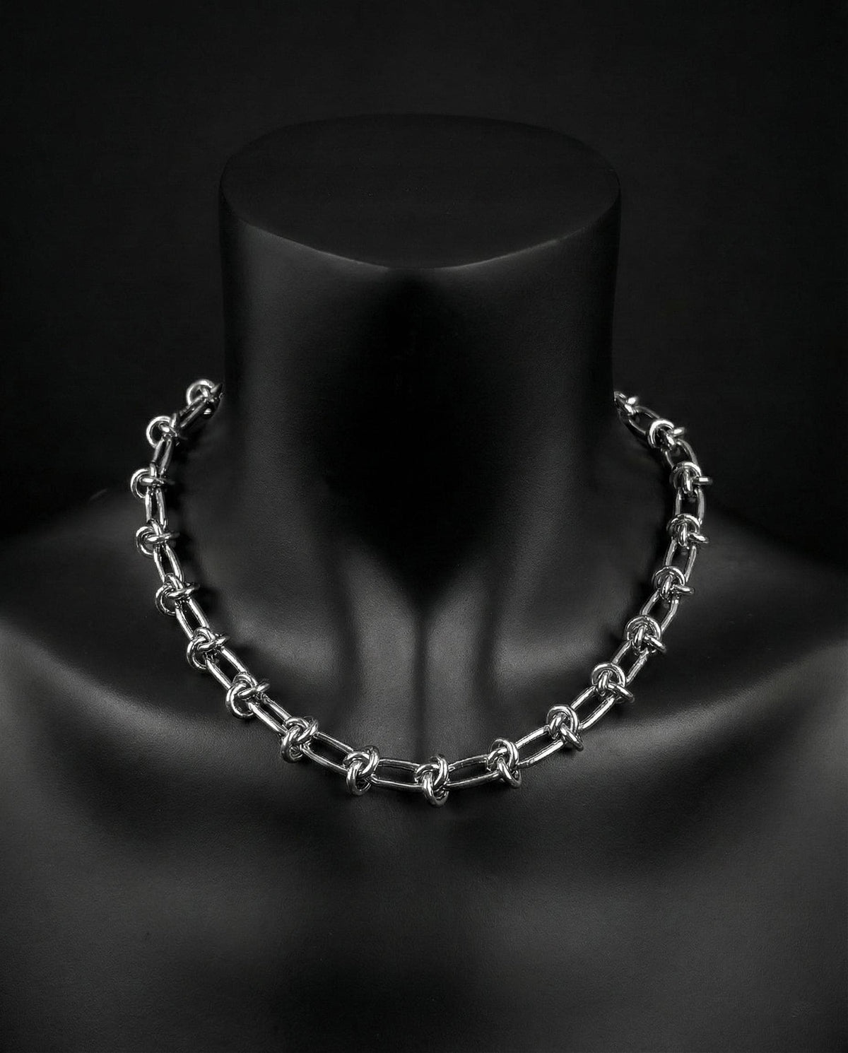 A silver stainless steel knotted chain necklace with a series of interlocking links. The necklace is displayed on a black mannequin, highlighting its design and texture. The chain is made of metal and has a polished finish.