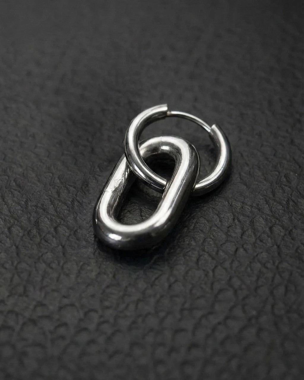 A single silver stainless steel metal earring with a distinctive design. It features a small hoop linked to a larger, elongated oval. The metal has a polished, reflective surface. The earring is set against a dark background.