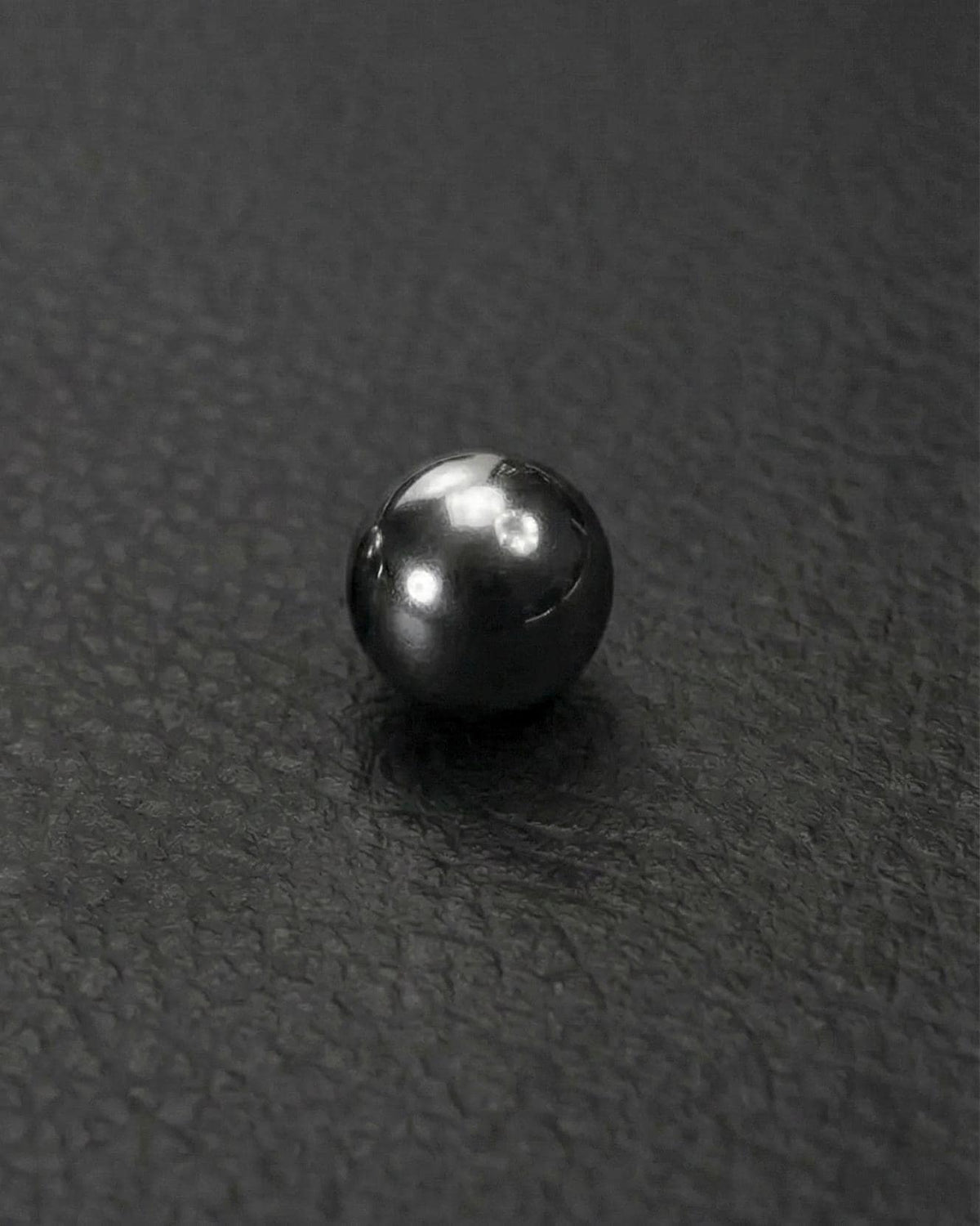A big, stainless steel, reflective, sphere, ball earring. The smooth, polished surface of the ball reflects light, creating bright highlights. The sphere is set against a textured, dark grey background.