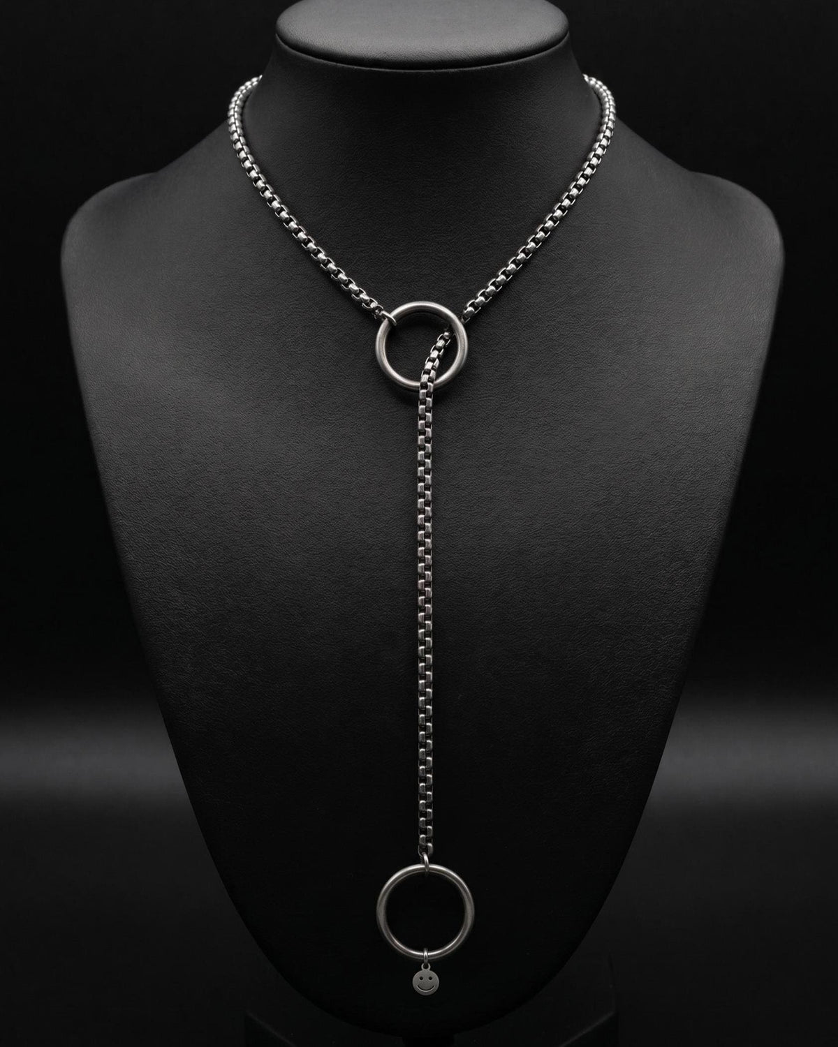 A silver-toned stainless steel lariat slip necklace featuring a box chain design. The chain passes through a circular ring at the centre, extending downwards to a second ring at the bottom. A tiny, circular charm with a smiley face is attached.