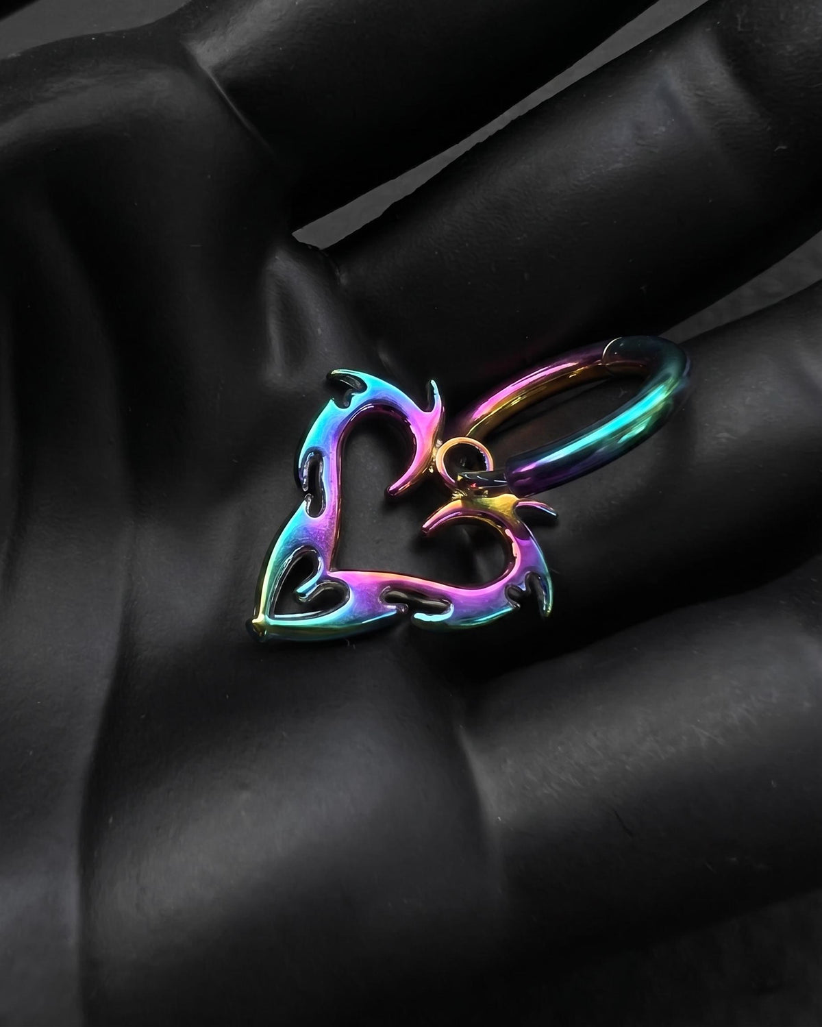 An iridescent rainbow-coloured stainless steel hoop earring featuring a dangling heart-shaped charm with flame-like cutouts. cyber sigilism style. The metal surface displays a gradient of blue, purple, pink, and yellow hues, creating a metallic, multi-coloured finish.
