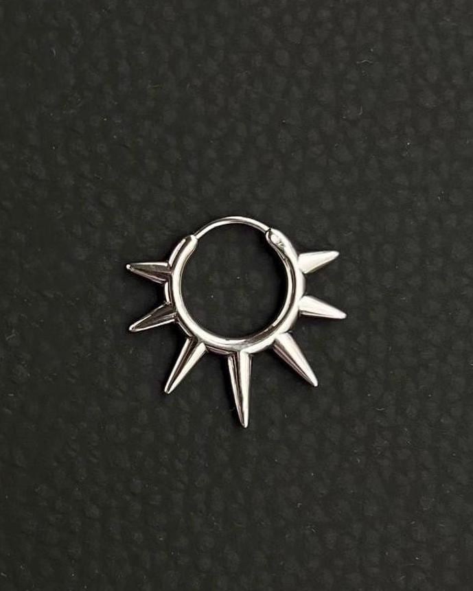 A silver-toned stainless steel circular hoop earring featuring a series of seven pointed spikes extending outwards from the outer edge of the ring. the earring looks like a symbolic sun, sun rays. The metal has a polished, reflective finish, and the design forms a sunburst or star-like shape.