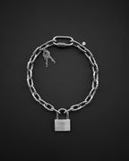 A silver-toned stainless steel chunky heavy thick industrial chain necklace featuring a rectangular padlock pendant at the bottom and a carabiner-style clasp at the top. Two small keys are attached to the chain links near the clasp, and a tiny circular charm is also present.