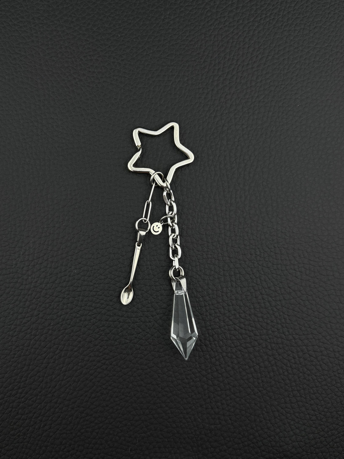 A silver-toned stainless steel keyring featuring a star-shaped ring, a small stainless steel metal spoon charm, a tiny circular smiley face charm, and a clear, faceted, diamond-shaped glass crystal pendant hanging from a short metal chain link.