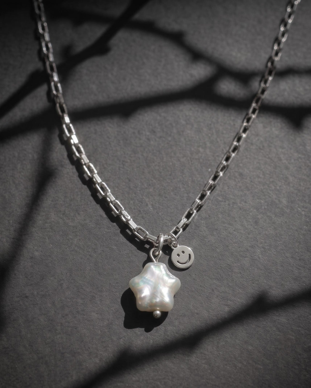 A silver-toned stainless steel chain necklace featuring a star-shaped real baroque white pearl pendant and a small circular charm with an engraved smiley face. The necklace has a box-style link chain design, and the pendant is attached with a simple silver jump ring.