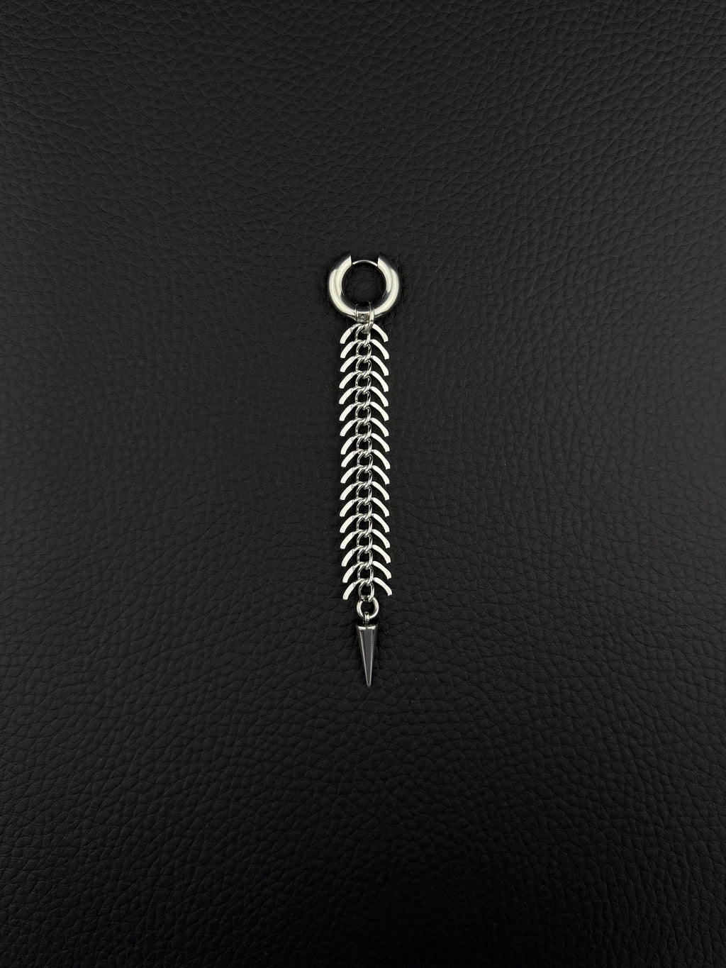 A silver-toned stainless steel hoop earring featuring a long, dangling chain composed of multiple curved, fishbone-like metal links. The chain terminates in a sharp, conical spike pendant, creating a vertical, articulated design with a polished metallic finish.