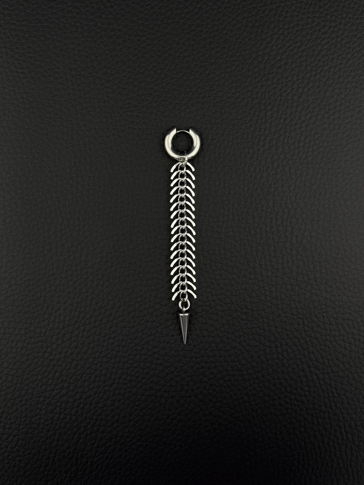 A silver-toned stainless steel hoop earring featuring a long, dangling chain composed of multiple curved, fishbone-like metal links. The chain terminates in a sharp, conical spike pendant, creating a vertical, articulated design with a polished metallic finish.