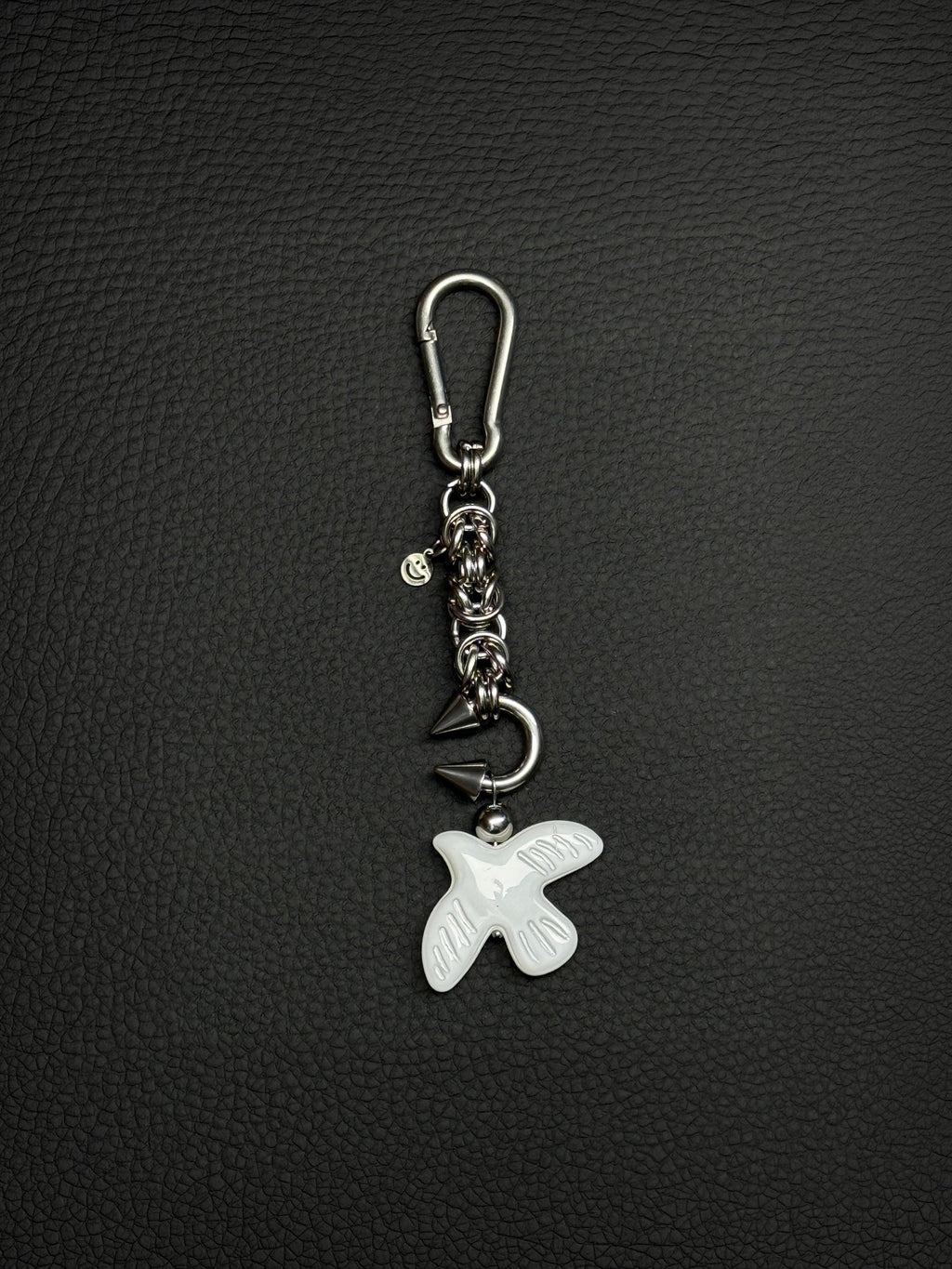 A silver-toned stainless steel metal keyring featuring a carabiner clip, a short chainmaille chain with circular links, a small smiley face charm, a curved metal barbel piercing earring with two pointed spikes, and a white glass peace dove bird-shaped pendant with etched wing details.