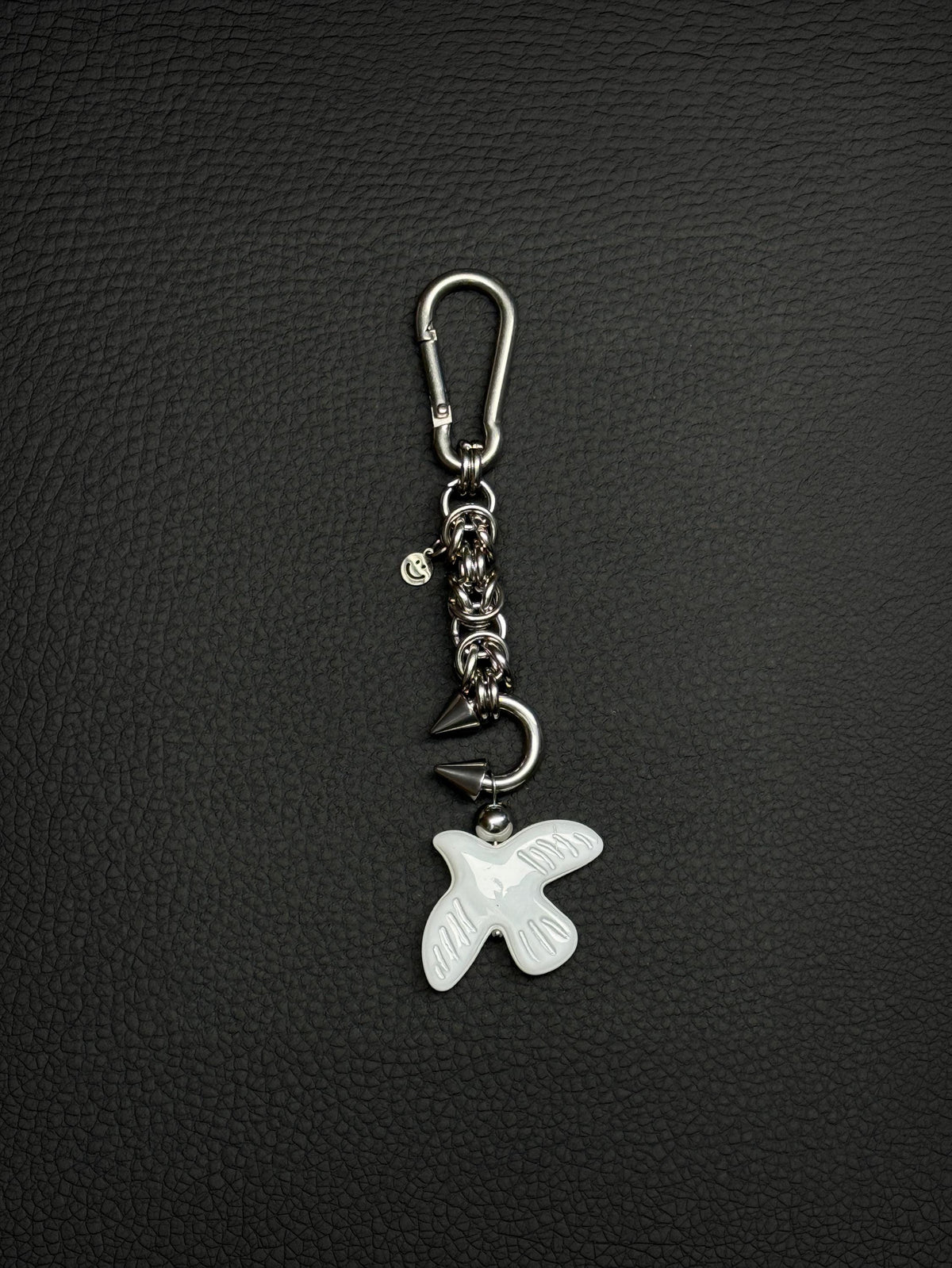 A silver-toned stainless steel metal keyring featuring a carabiner clip, a short chainmaille chain with circular links, a small smiley face charm, a curved metal barbel piercing earring with two pointed spikes, and a white glass peace dove bird-shaped pendant with etched wing details.