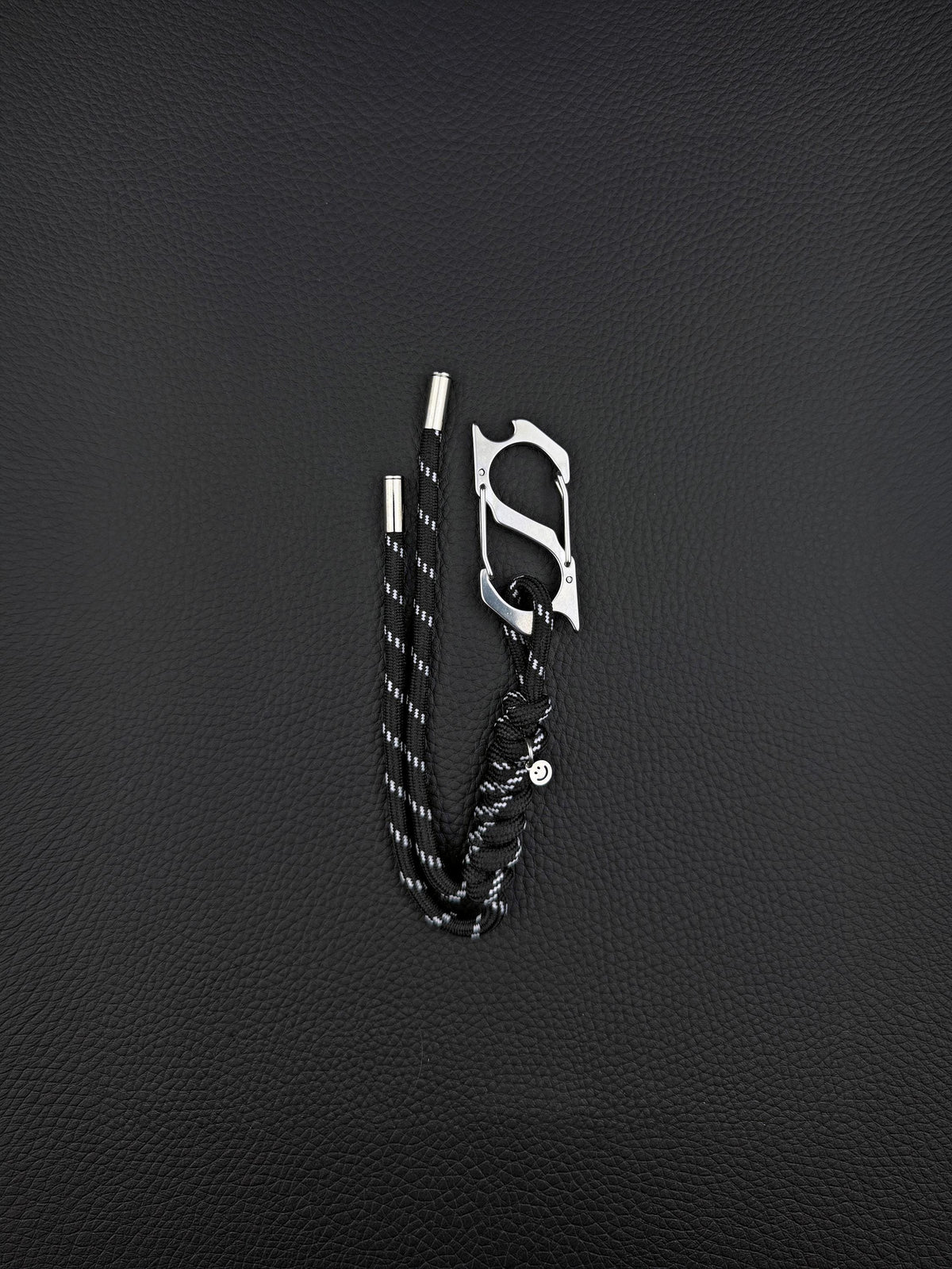 A black paracord keyring featuring a silver-toned stainless steel metal carabiner with an integrated bottle opener. The cord is woven with reflective white threads and finished with two silver metal aglets at the ends and a small circular charm. cyber sigilism style