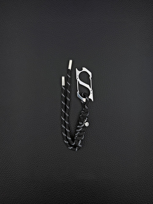 A black paracord keyring featuring a silver-toned stainless steel metal carabiner with an integrated bottle opener. The cord is woven with reflective white threads and finished with two silver metal aglets at the ends and a small circular charm. cyber sigilism style