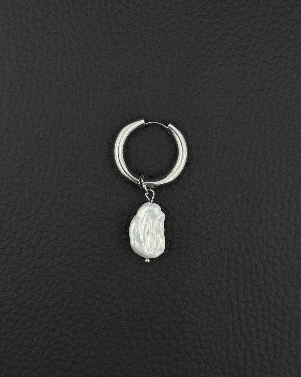 A silver-coloured stainless steel metal hoop earring featuring a single, irregular, white baroque pearl charm suspended from the bottom. The hoop has a smooth, polished finish and a hinged closure mechanism, while the pearl displays a textured surface.