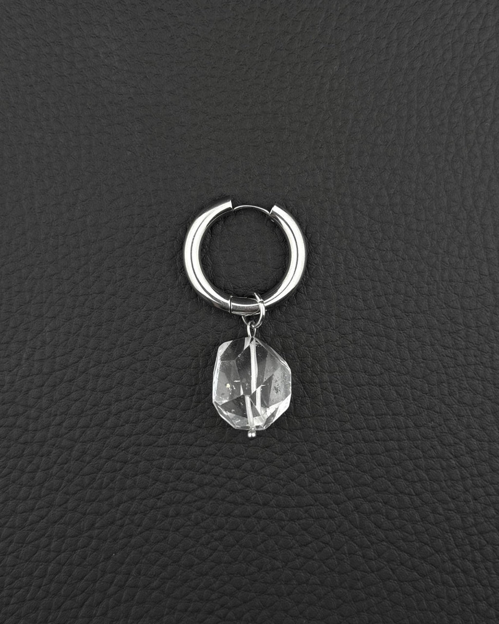 A silver-toned stainless steel  metal hoop earring featuring a single clear, faceted, irregular-shaped quartz crystal pendant hanging from the bottom. The earring has a smooth, polished finish and a hinged closure mechanism at the top of the circular hoop.