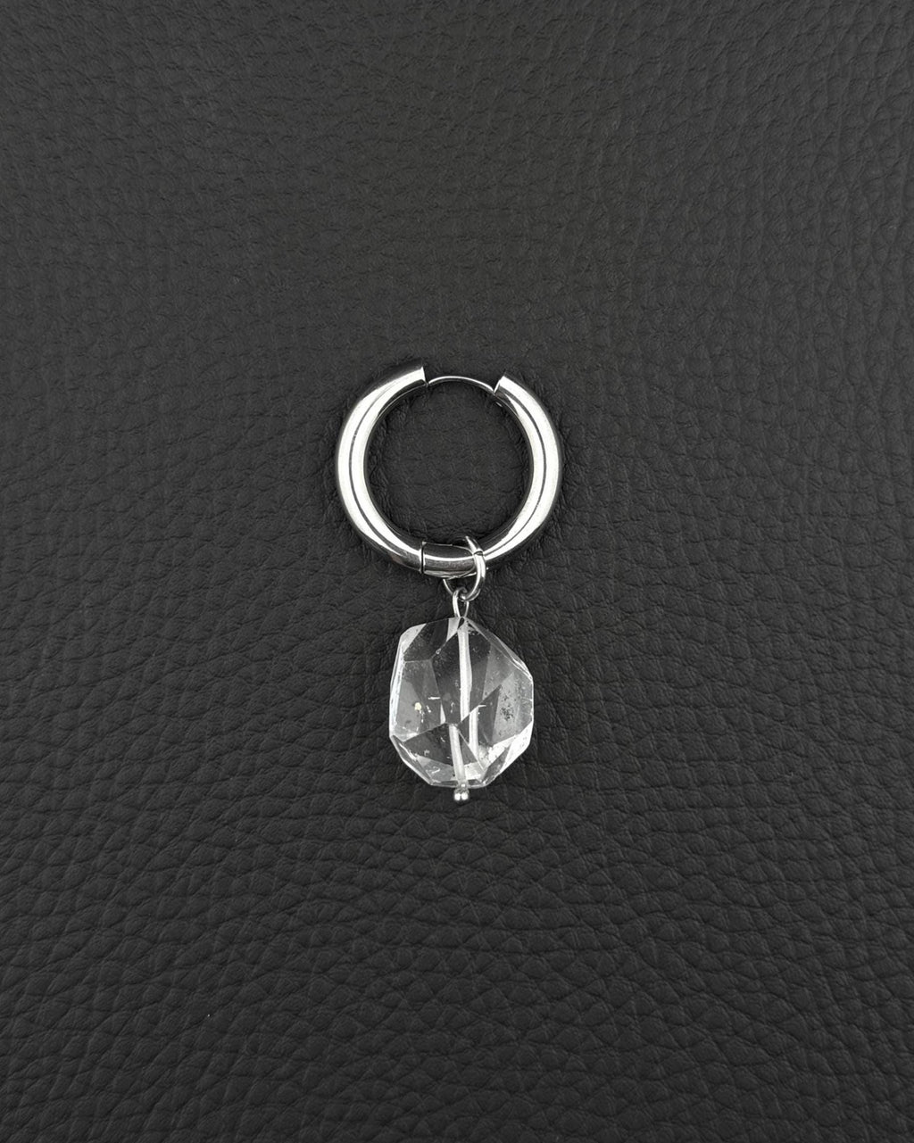 A silver stainless steel hoop earring featuring a single clear, faceted, irregular-shaped quartz crystal pendant hanging from the bottom. The earring has a smooth, polished finish and a hinged closure mechanism at the top of the circular hoop.