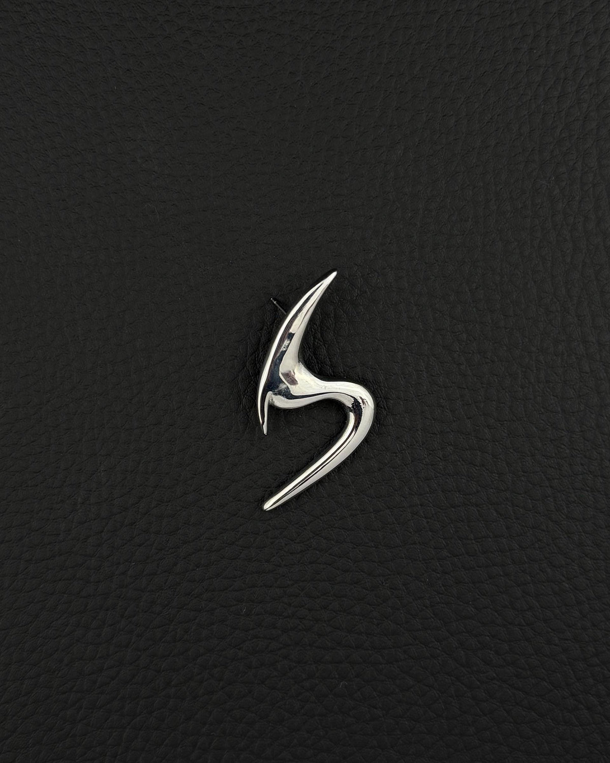 A polished silver-tone stainless steel brooch featuring a sleek, abstract, curved design that resembles a stylised letter S. The piece has a smooth, reflective surface with sharp, pointed ends and a fluid, organic shape that tapers at both extremities.