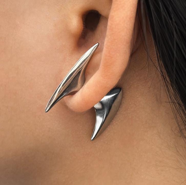A silver-toned stainless steel ear cuff featuring two distinct, smooth, pointed, and curved metal spikes. The larger, elongated spike rests along the upper outer edge of the ear, while the smaller, tapered spike sits below the earlobe, creating a sleek look.