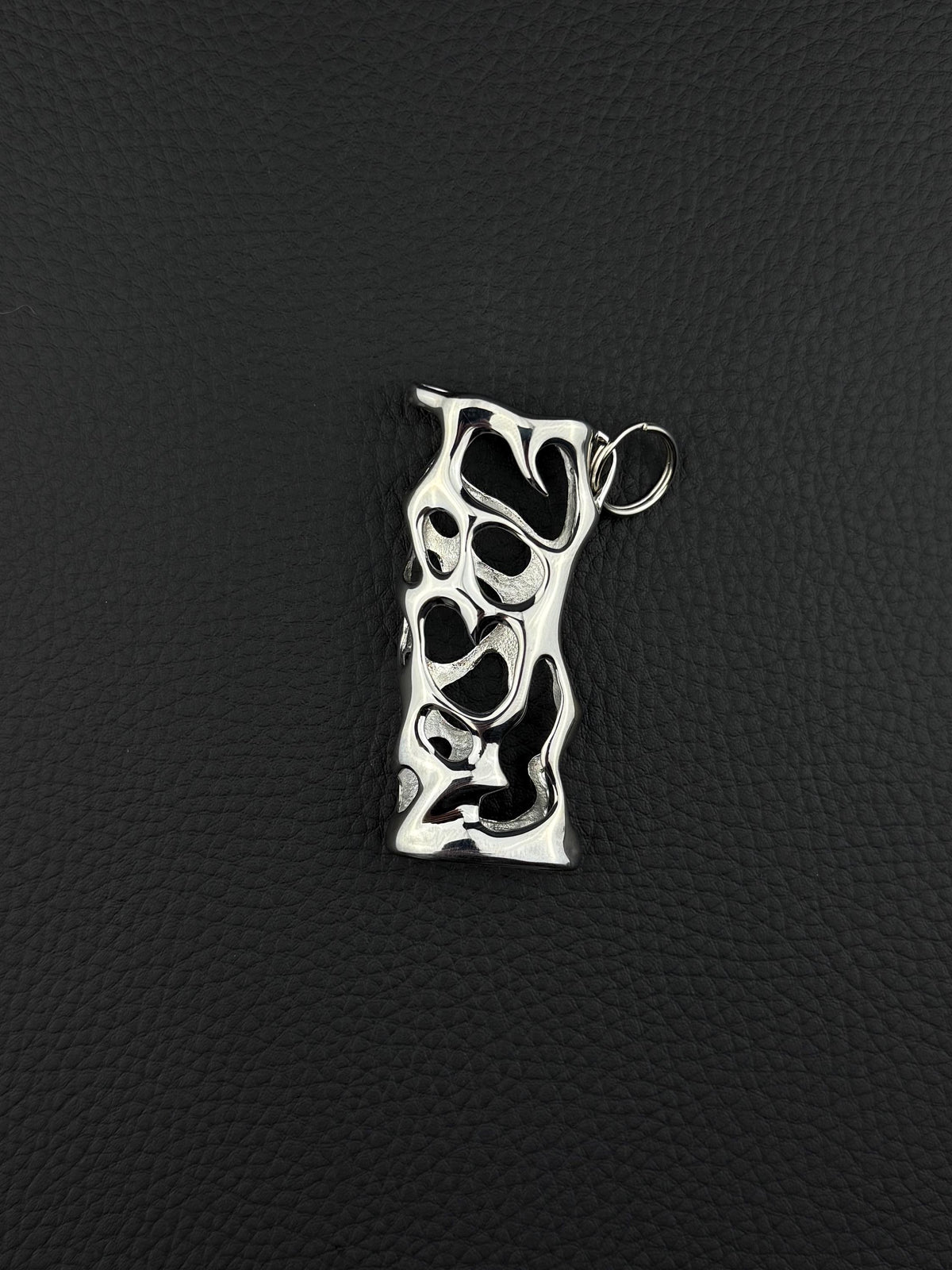 A silver-toned stainless steel pendant lighter sleeve featuring an elongated, irregular rectangular shape with a complex, organic cutout pattern throughout its surface. The metal has a polished, reflective finish and includes a small circular jump ring attached at the top. cyber sigilism style