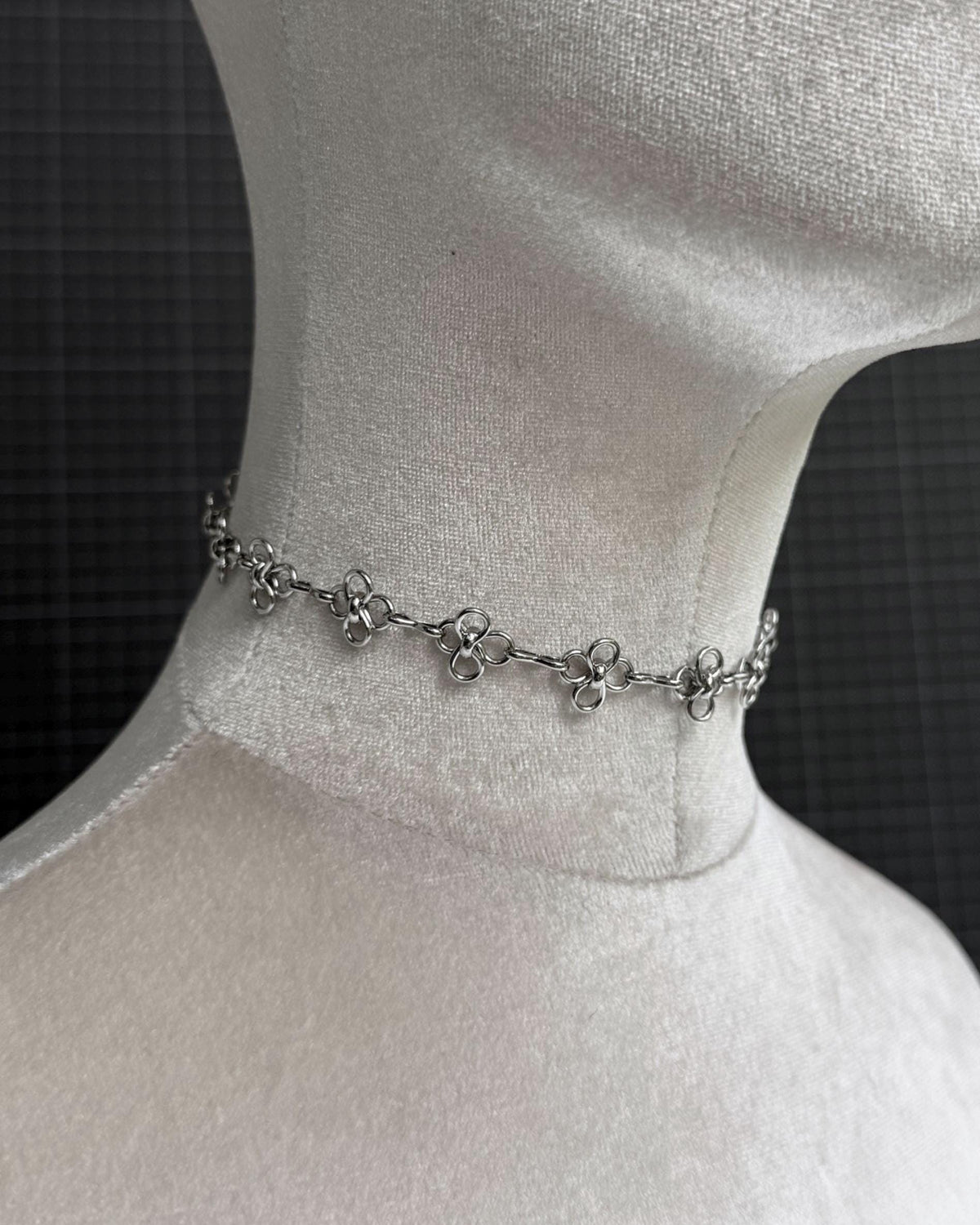 A silver-toned stainless steel tattoo choker necklace featuring a repeating pattern of small, four-petaled flower-shaped links connected by thin circular rings. The chain sits snugly around the neck of a white fabric mannequin display form.