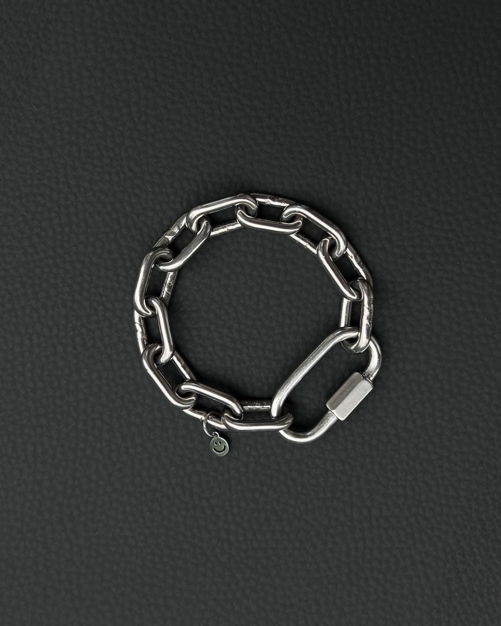 A silver-toned stainless steel chunky chain bracelet composed of thick, interlocking rectangular links. The bracelet features a large, elongated carabiner-style clasp and a small, circular charm engraved with a simple smiley face icon.