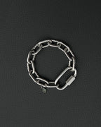 A silver-toned stainless steel chunky chain bracelet composed of thick, interlocking rectangular links. The bracelet features a large, elongated carabiner-style clasp and a small, circular charm engraved with a simple smiley face icon.