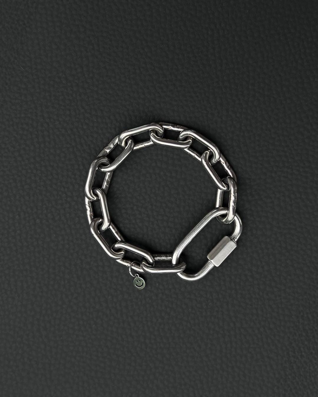 A silver-toned stainless steel chunky chain bracelet composed of thick, interlocking rectangular links. The bracelet features a large, elongated carabiner-style clasp and a small, circular charm engraved with a simple smiley face icon.