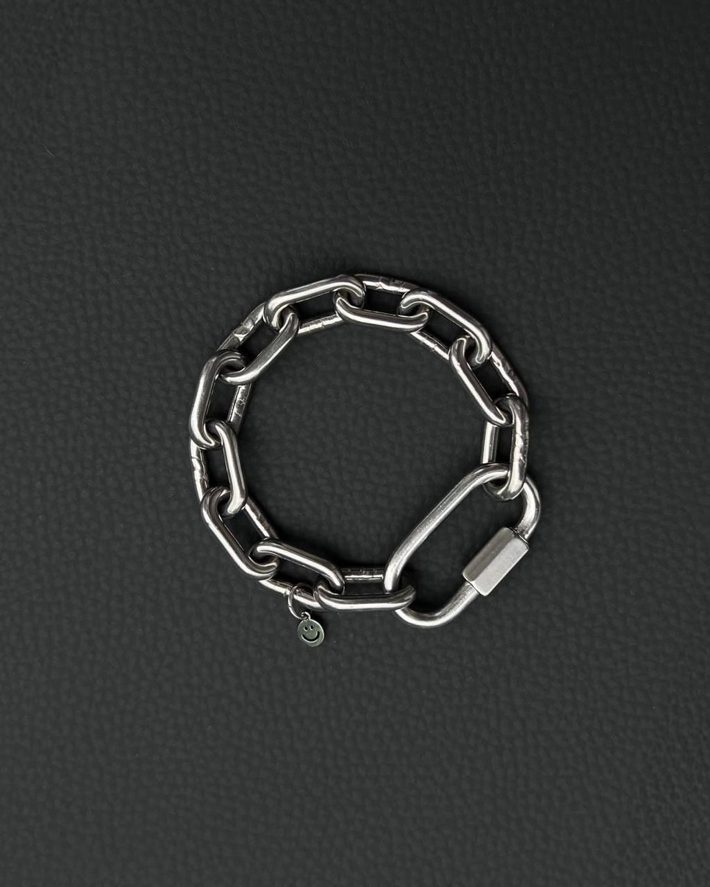 A silver-toned stainless steel chunky chain bracelet composed of thick, interlocking rectangular links. The bracelet features a large, elongated carabiner-style clasp and a small, circular charm engraved with a simple smiley face icon.