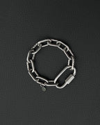 A silver-toned stainless steel chunky chain bracelet composed of thick, interlocking rectangular links. The bracelet features a large, elongated carabiner-style clasp and a small, circular charm engraved with a simple smiley face icon.