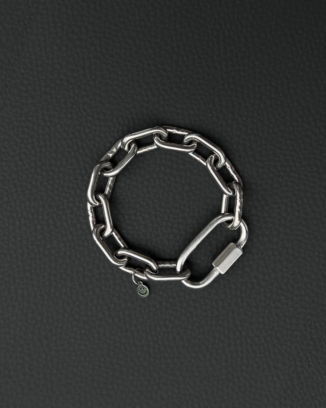 A silver-toned stainless steel chunky chain bracelet composed of thick, interlocking rectangular links. The bracelet features a large, elongated carabiner-style clasp and a small, circular charm engraved with a simple smiley face icon.