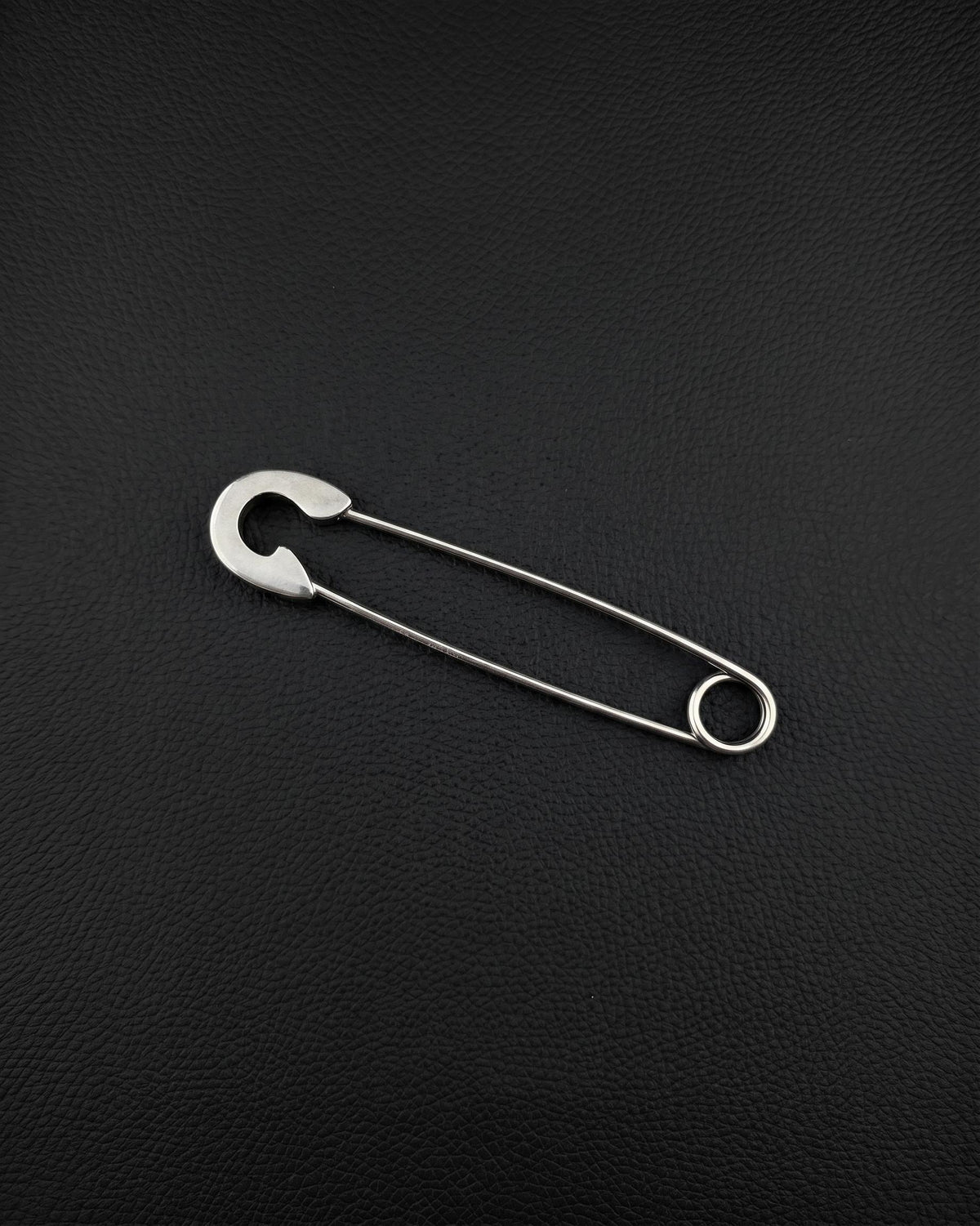 A silver-coloured stainless steel safety pin earring featuring a classic U-shaped coil at one end and a protective clasp mechanism at the other. The pin is constructed from a single piece of wire, showcasing a smooth, polished finish and a functional design.