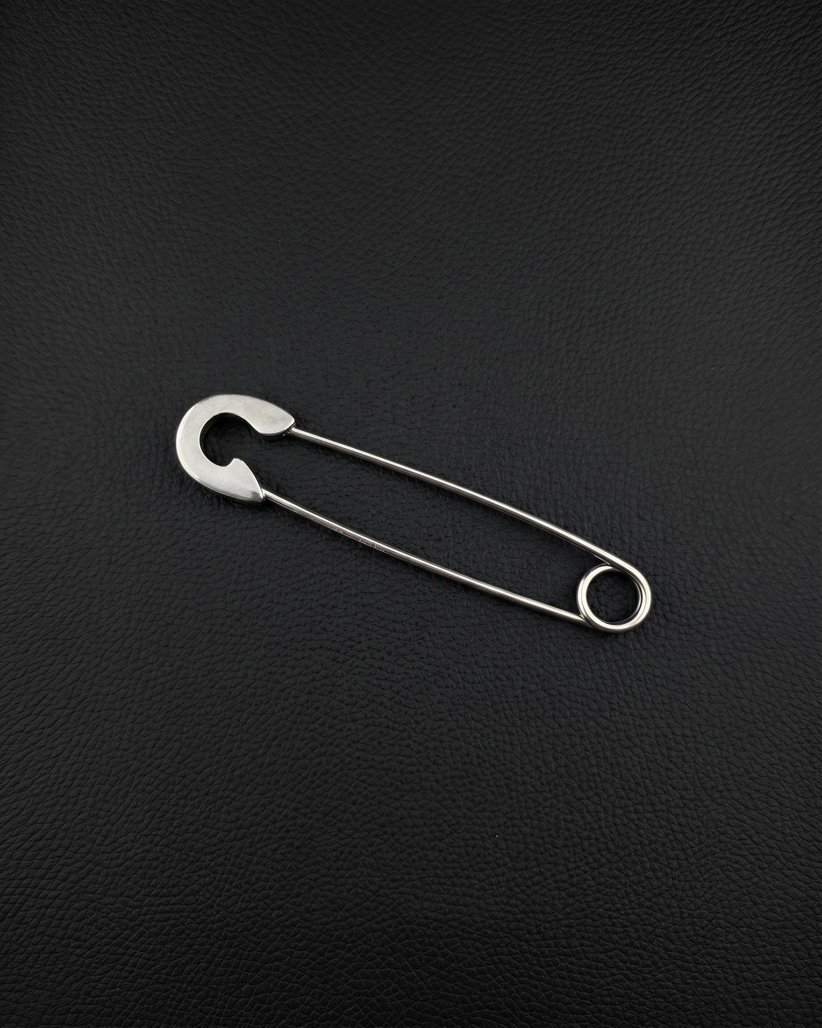A silver-coloured stainless steel safety pin earring featuring a classic U-shaped coil at one end and a protective clasp mechanism at the other. The pin is constructed from a single piece of wire, showcasing a smooth, polished finish and a functional design.