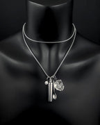 A silver-toned double-layered chain necklace featuring three hanging charms: a small cylindrical metal capsule, a tiny spoon, and a clear, faceted quartz stone. A small circular charm with a smiley face is also attached.